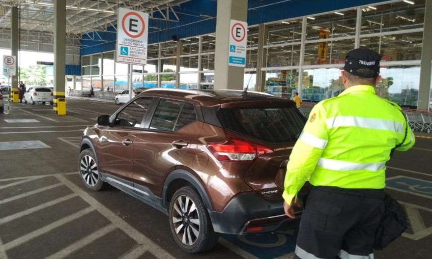 Fiscal de trânsito em vaga para PcD ao lado de carro marrom em estacionamento.
