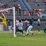 Goleiro pula para defender a bola em jogo de futebol, com jogadores em campo e torcida ao fundo.