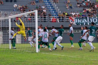 Goleiro pula para defender a bola em jogo de futebol, com jogadores em campo e torcida ao fundo.