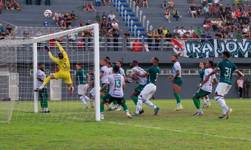 Goleiro pula para defender a bola em jogo de futebol, com jogadores em campo e torcida ao fundo.