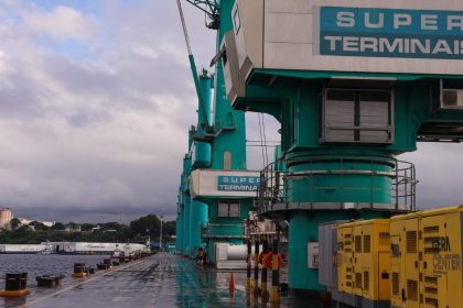 Guindastes de carga no porto com o céu nublado ao fundo. O letreiro diz 'SUPER TERMINAIS'.