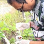 Homem com luvas e vestindo uniforme da Amazônia manipulando substância próximo à vegetação, em combate à dengue.