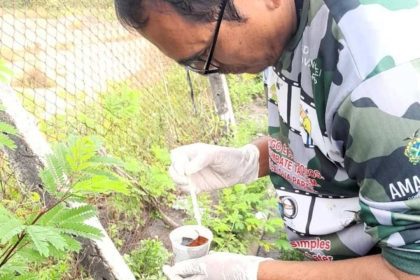 Homem com luvas e vestindo uniforme da Amazônia manipulando substância próximo à vegetação, em combate à dengue.