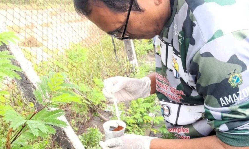 Homem com luvas e vestindo uniforme da Amazônia manipulando substância próximo à vegetação, em combate à dengue.