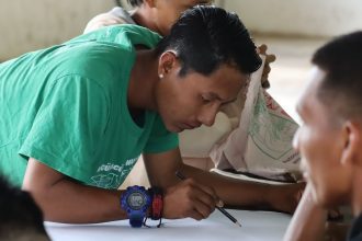 Homem desenhando em papel, com colegas ao redor.