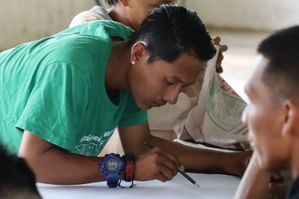 Homem desenhando em papel, com colegas ao redor.