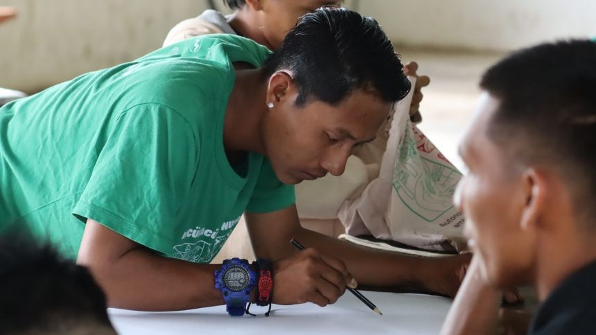 Homem desenhando em papel, com colegas ao redor.