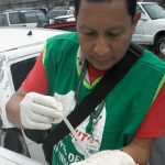 Homem em vestimenta de proteção coleta amostra em carro danificado, possivelmente para investigação de dengue.
