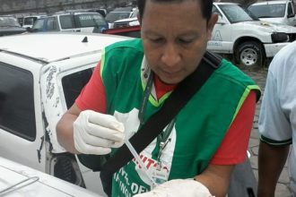 Homem em vestimenta de proteção coleta amostra em carro danificado, possivelmente para investigação de dengue.