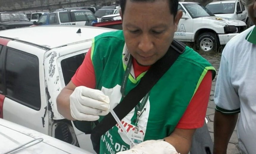Homem em vestimenta de proteção coleta amostra em carro danificado, possivelmente para investigação de dengue.
