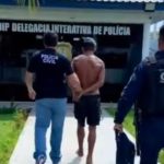 Homem sendo escoltado por policiais em frente à delegacia de polícia.