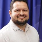 Prefeito sorrindo com barba e camisa clara em frente a cortina azul.