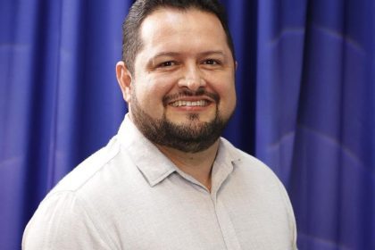 Prefeito sorrindo com barba e camisa clara em frente a cortina azul.