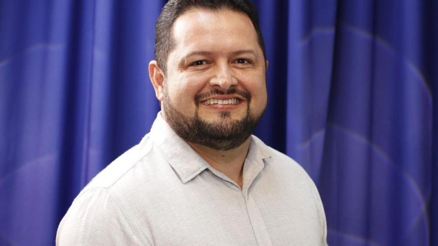 Prefeito sorrindo com barba e camisa clara em frente a cortina azul.