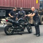 Homens investigam uma moto preta em uma via, com caminhão ao fundo. Possível cena de acidente envolvendo um militar.