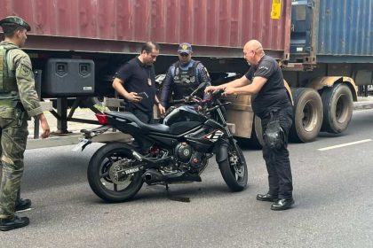Homens investigam uma moto preta em uma via, com caminhão ao fundo. Possível cena de acidente envolvendo um militar.