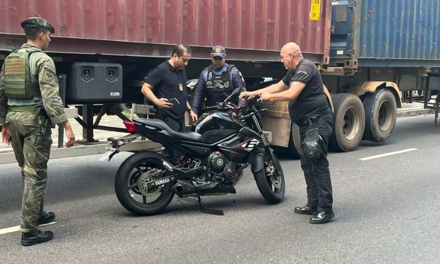 Homens investigam uma moto preta em uma via, com caminhão ao fundo. Possível cena de acidente envolvendo um militar.