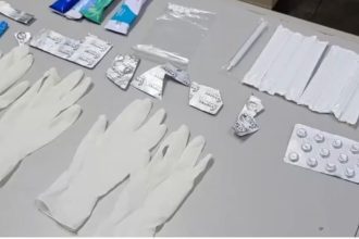 Itens médicos, como luvas, embalagens de remédios e seringas, dispostos sobre uma superfície.