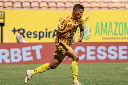 Jogador de futebol comemorando gol em campo, vestindo uniforme amarelo e preto.