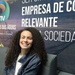Valentina Marques sorrindo sentada em um sofá, com cabelo cacheado e jaqueta jeans, em frente a um banner.