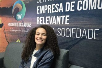 Valentina Marques sorrindo sentada em um sofá, com cabelo cacheado e jaqueta jeans, em frente a um banner.