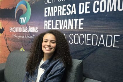 Valentina Marques sorrindo sentada em um sofá, com cabelo cacheado e jaqueta jeans, em frente a um banner.