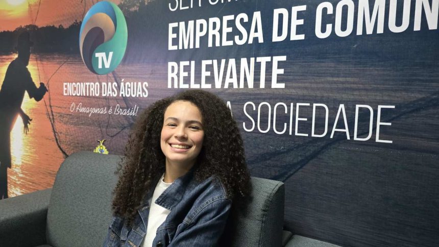 Valentina Marques sorrindo sentada em um sofá, com cabelo cacheado e jaqueta jeans, em frente a um banner.
