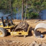 Máquina compactadora amarela em área de construção próxima a rio, possivelmente em área de preservação.