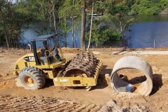 Máquina compactadora amarela em área de construção próxima a rio, possivelmente em área de preservação.
