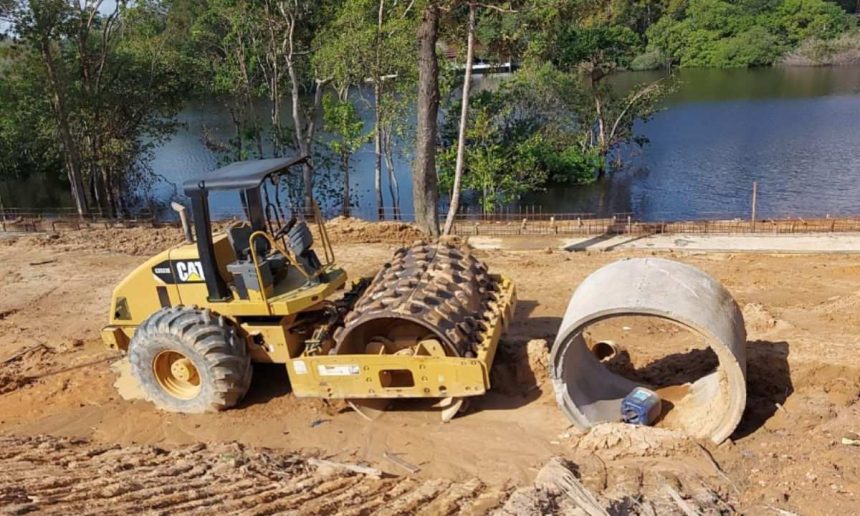 Máquina compactadora amarela em área de construção próxima a rio, possivelmente em área de preservação.