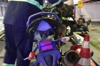 Motocicleta sendo removida por bombeiros após acidente, com cones de sinalização e pessoas no local.