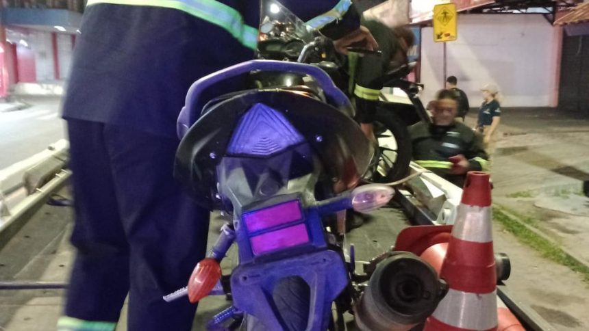 Motocicleta sendo removida por bombeiros após acidente, com cones de sinalização e pessoas no local.