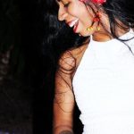 Mulher sorrindo com flores no cabelo e top branco, em evento de música latina.