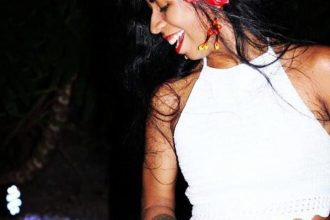 Mulher sorrindo com flores no cabelo e top branco, em evento de música latina.