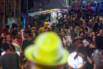 Multidão em festa na rua Barroso, com barracas e luzes coloridas à noite.