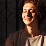 Nando Montenegro sorrindo com os olhos fechados, em um ensaio com luz solar, com grade de fundo