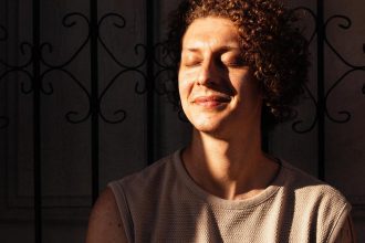 Nando Montenegro sorrindo com os olhos fechados, em um ensaio com luz solar, com grade de fundo