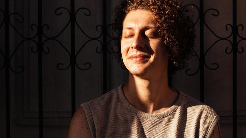 Nando Montenegro sorrindo com os olhos fechados, em um ensaio com luz solar, com grade de fundo