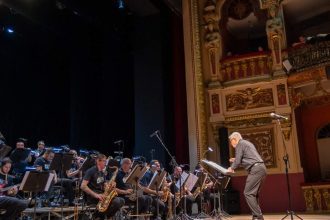 Orquestra da Amazonas Band em palco com maestro e músicos tocando instrumentos de sopro e guitarra. Público na plateia.