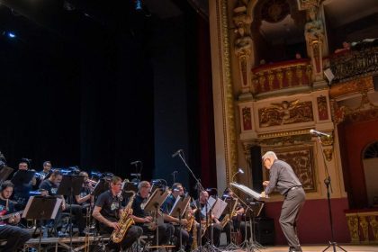 Orquestra da Amazonas Band em palco com maestro e músicos tocando instrumentos de sopro e guitarra. Público na plateia.