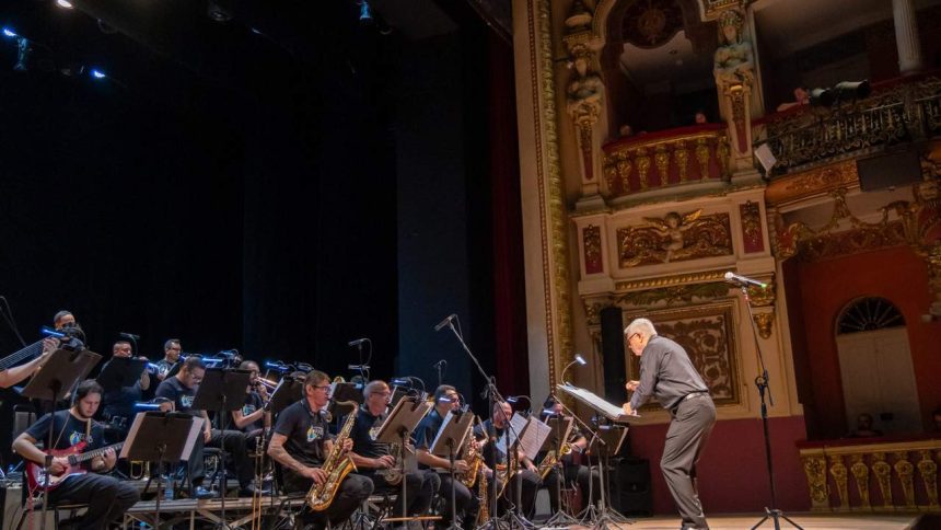 Orquestra da Amazonas Band em palco com maestro e músicos tocando instrumentos de sopro e guitarra. Público na plateia.