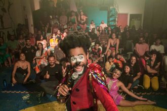 Palhaço se apresentando em um palco, com grande público ao fundo.