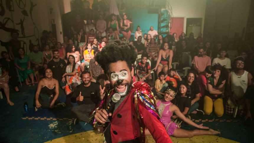 Palhaço se apresentando em um palco, com grande público ao fundo.