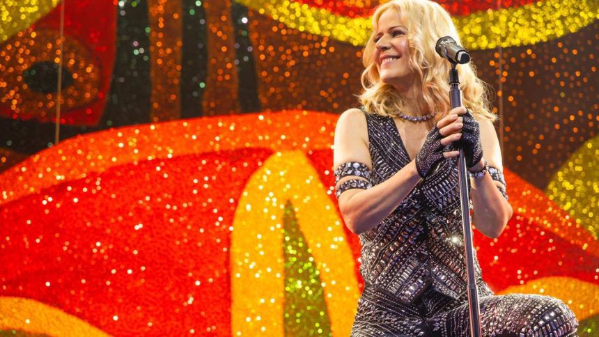 Paula Toller no palco, sorrindo, com figurino brilhante, segurando o microfone, em show.