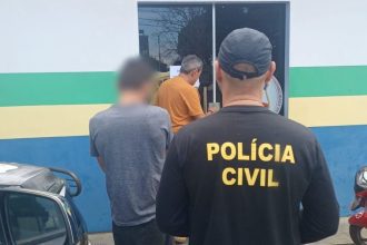 Policial civil com uniforme preto, em frente a uma delegacia, com presos.