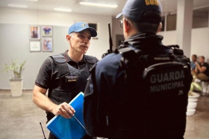 Policial conversa com colega em operação policial. Um deles usa colete e boné.