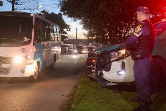 Policial em frente a viatura e ônibus, em celebração a segurança no transporte de trabalhadores em Manaus.