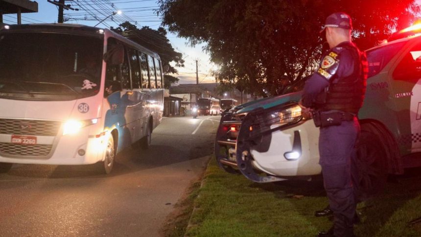 Policial em frente a viatura e ônibus, em celebração a segurança no transporte de trabalhadores em Manaus.