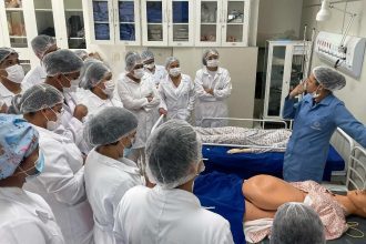 Professora de enfermagem demonstra em manequim para turma em sala de aula.