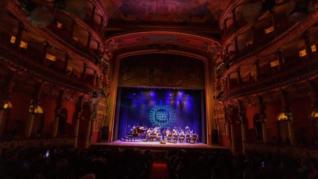 Show de jazz no palco de um teatro antigo, com banda e público presente.